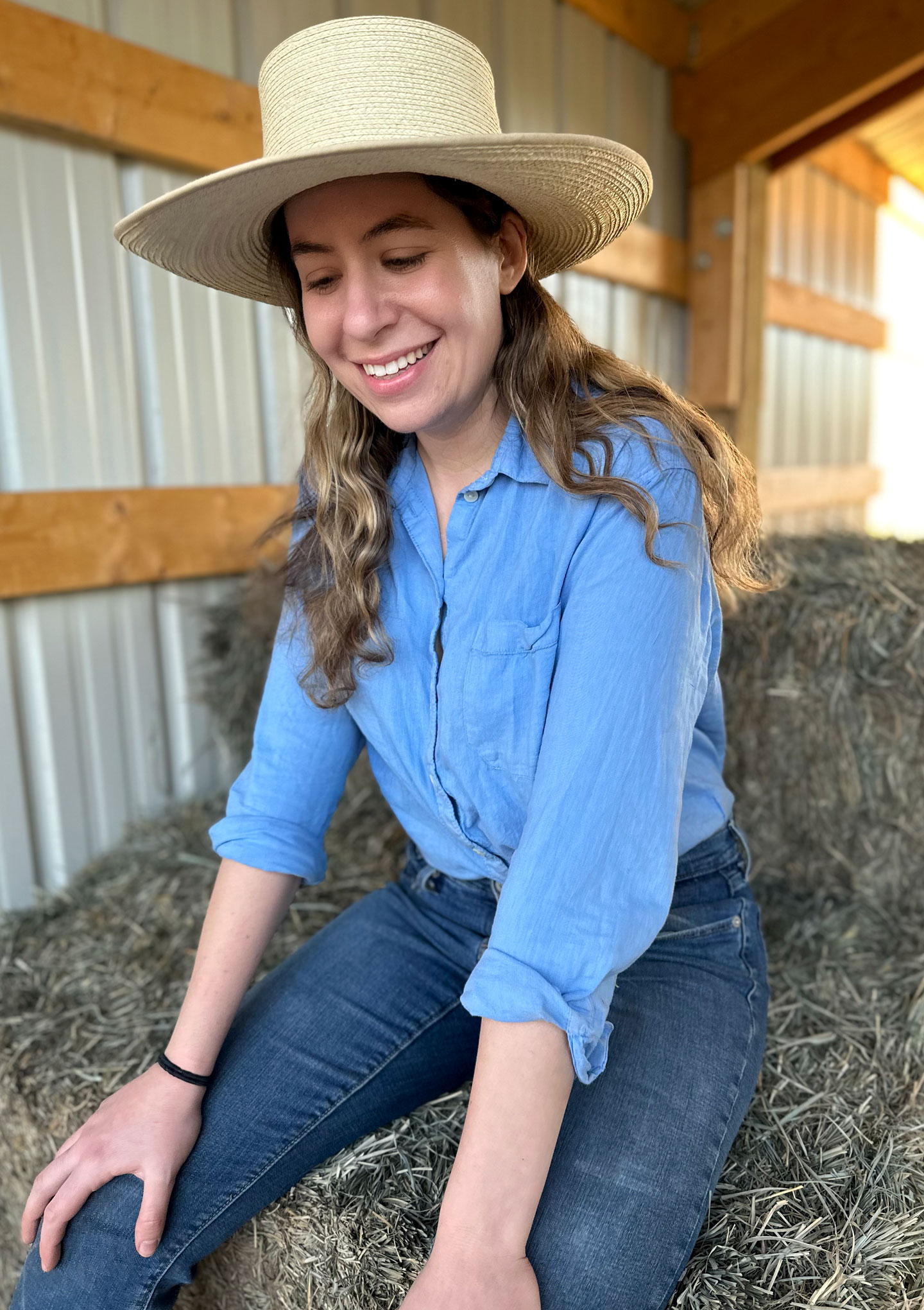 Rebecca is sitting on a bale of orchard grass in a barn